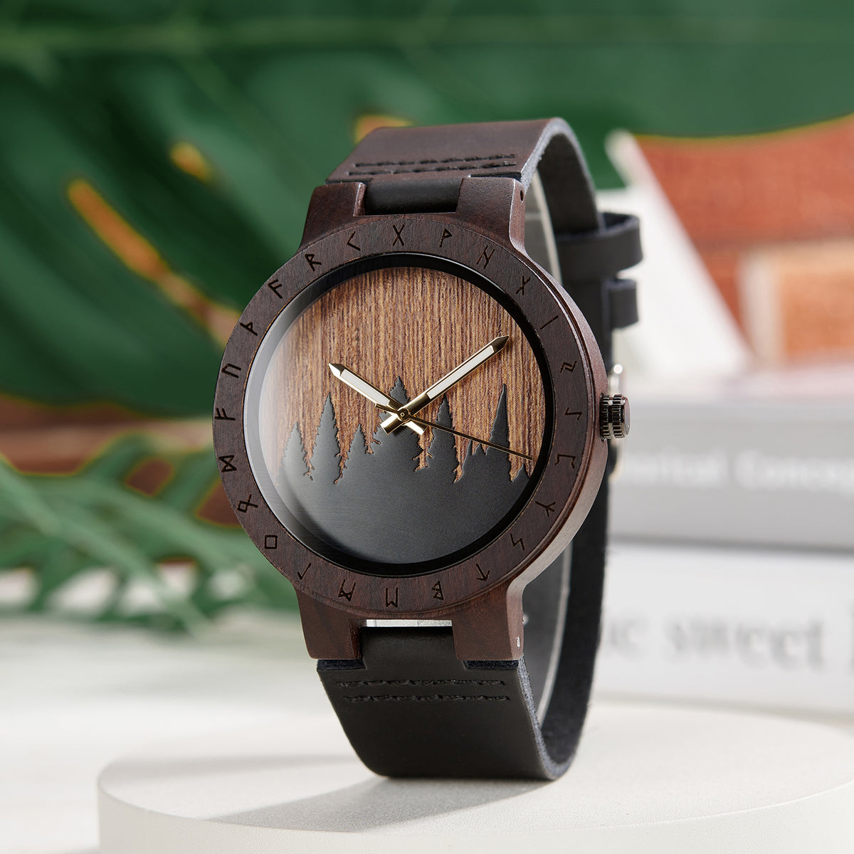 A unique dark wooden watch with a runic symbol bezel, forest silhouette dial, and black leather band, featuring gold-tone hands, displayed against a blurred background of green foliage and books.