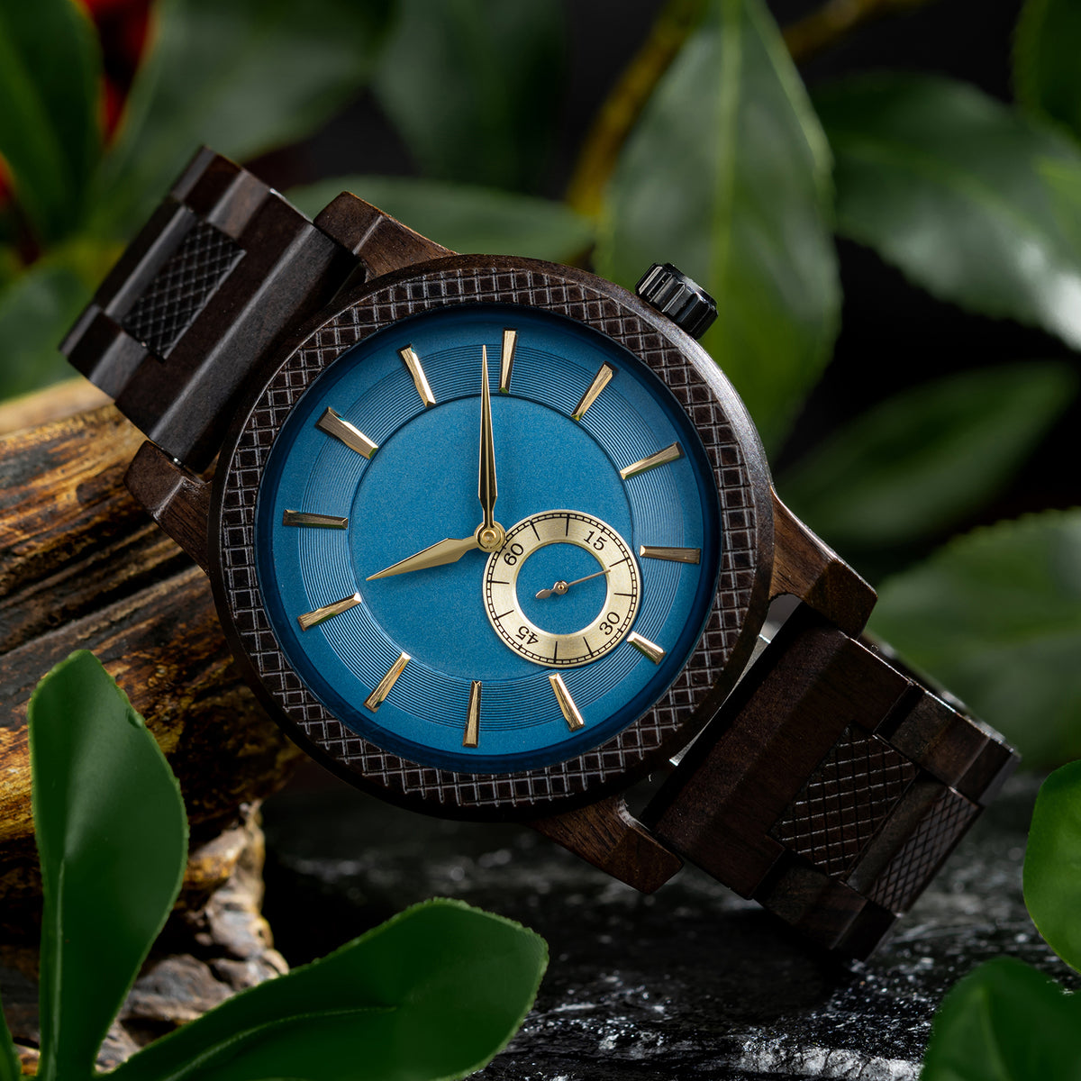 A stylish men's wooden watch featuring a blue dial, sub-dial, textured bezel, and gold-accented details, presented against a backdrop of rustic wood and green foliage to highlight its sophisticated and natural aesthetic.