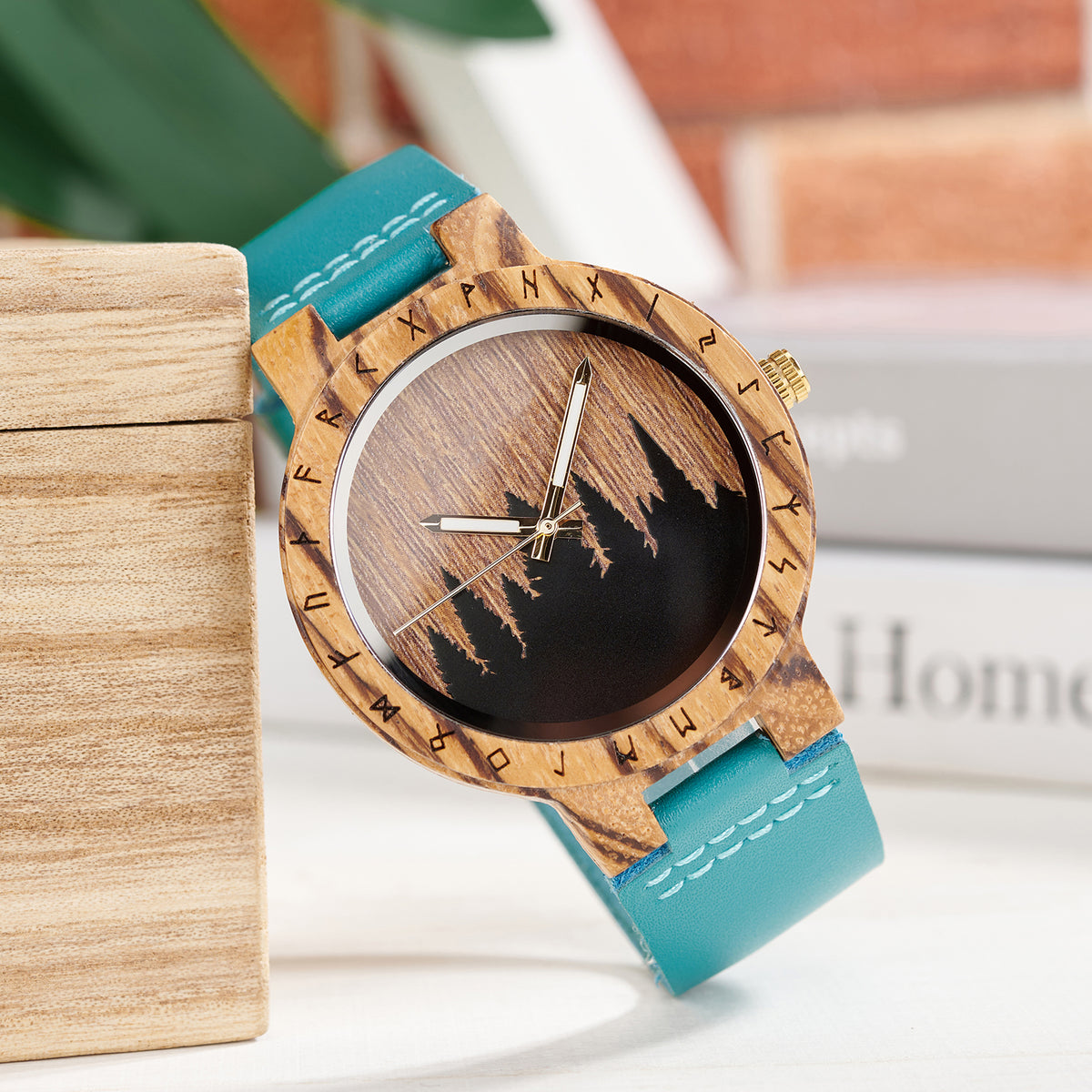 A unique wooden watch featuring a runic symbol bezel, mountain-themed wood dial, and turquoise leather band, placed next to a wooden box with a blurred background of greenery and books