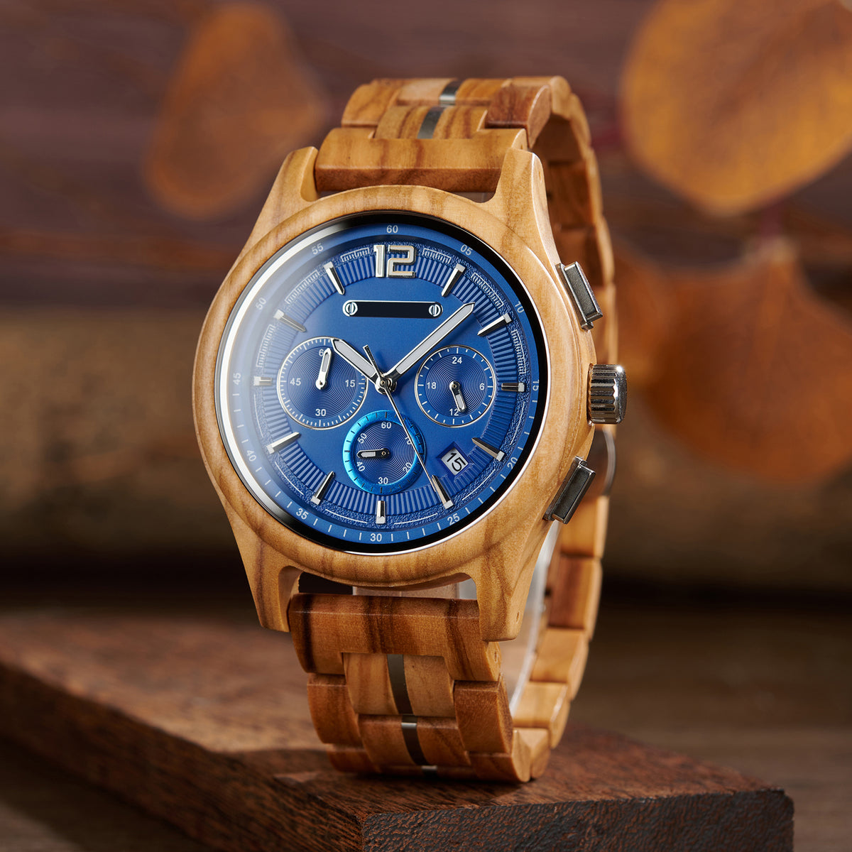 A wooden chronograph watch with a blue dial, multiple sub-dials, date "10" display, and a strap made of wooden links with metal accents, positioned on a dark wooden block against a background with blurred autumn leaves.