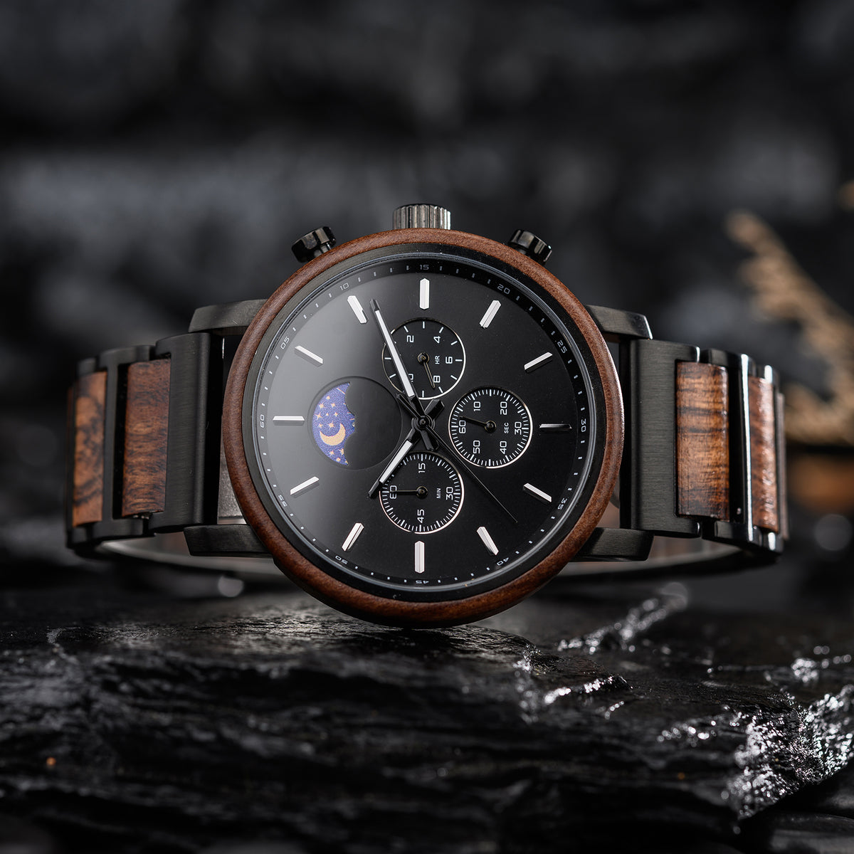 A sophisticated black multi-function watch featuring a moon phase complication, with a band combining wood and black metal, placed on dark, textured rocks.