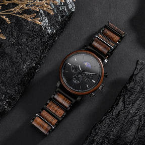 A sophisticated chronograph watch with a wooden bezel, black metal and wood combined band, featuring a moon phase complication on a black dial, placed on dark rocks with dried plants in the background.