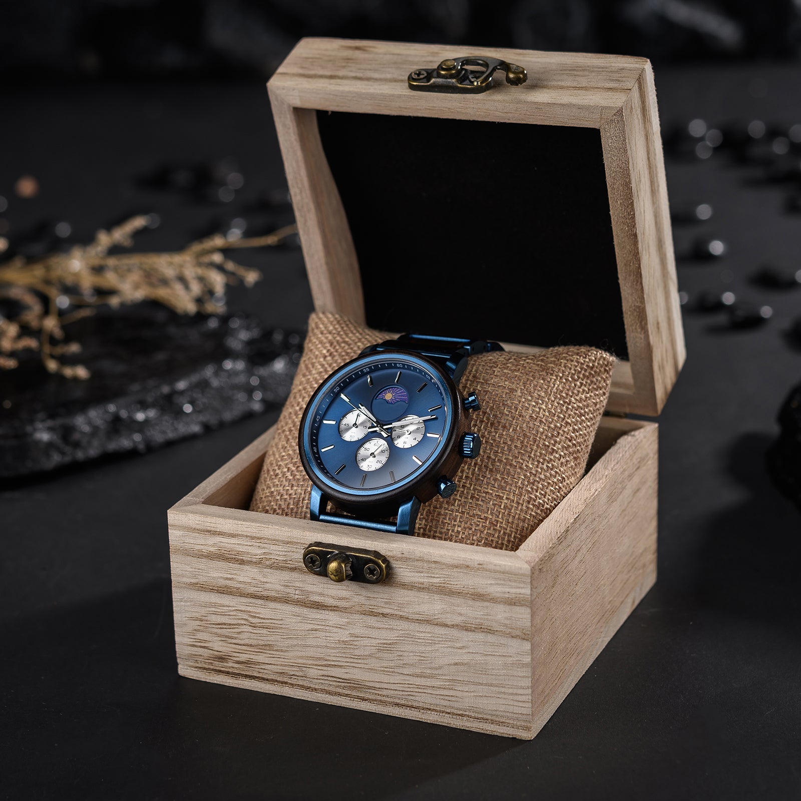 A stylish blue multi-function watch with moon phase complication, presented in an open light wooden box with a burlap pillow, set against a dark, decorative background.
