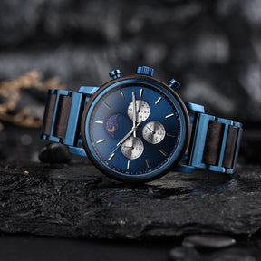 A stylish blue chronograph watch with a combination of wood and metal band, featuring a moon phase complication on a blue dial, placed on dark rocks.