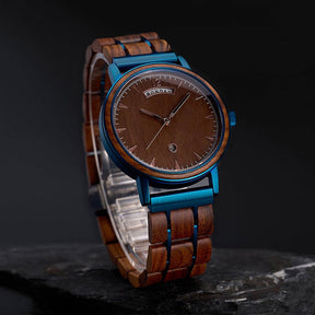 A wristwatch featuring a brown wooden dial showing "MONDAY", with a strap made of wooden links and blue metal accents, positioned on stacked black rocks against a dark background.