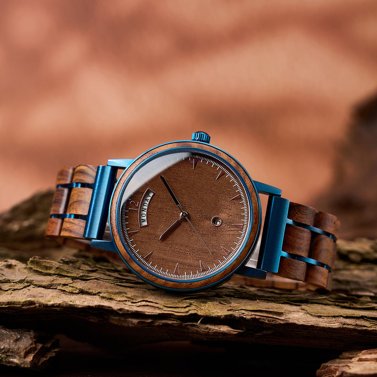 A wristwatch featuring a brown wooden dial showing "MONDAY" for the day, with a strap made of wooden links and blue metal accents, placed on textured tree bark against a warm, blurred background.