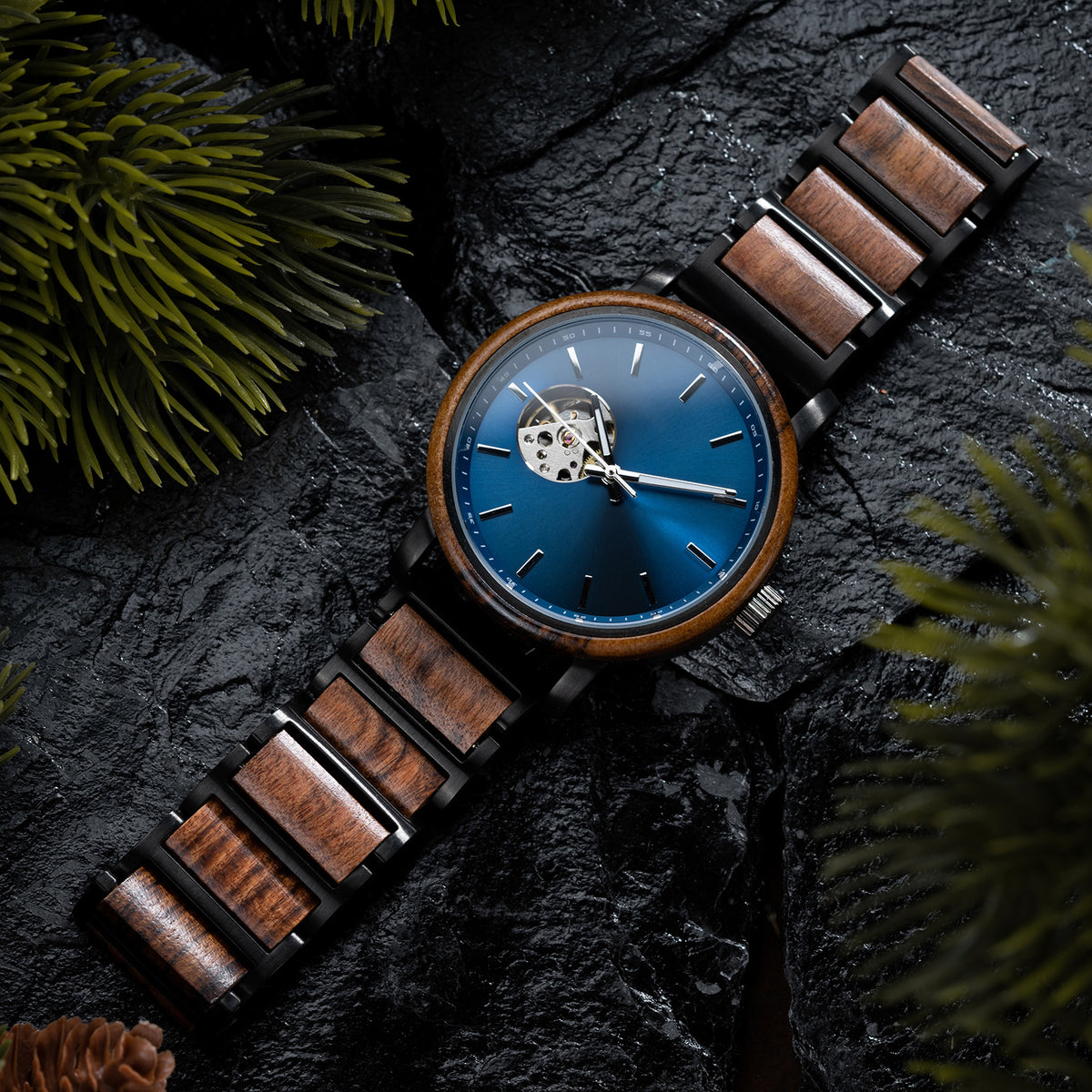 A wristwatch with a blue dial, skeleton movement window, and a band combining wood and black metal, placed on dark rocky surface with green pine foliage and a pine cone.
