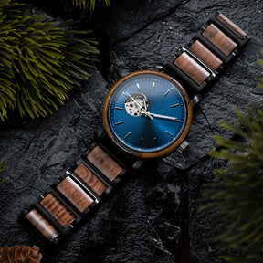 A wristwatch with a blue dial, skeleton movement window, and a band combining wood and black metal, placed on dark rocky surface with green pine foliage and a pine cone.
