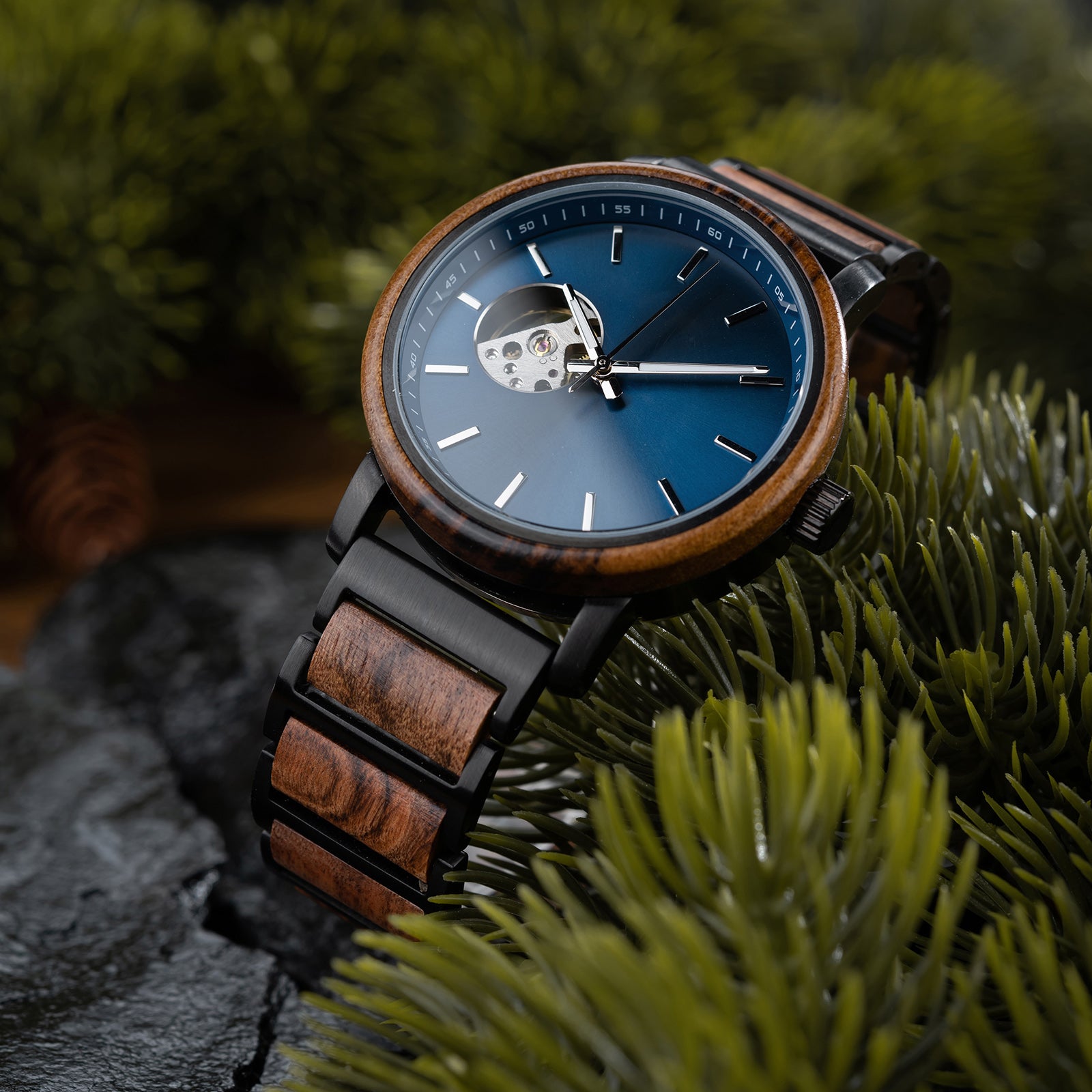 A wristwatch with a blue dial, skeleton movement window, and a band combining wood and black metal, placed on green pine foliage with dark rocky surface in the background.