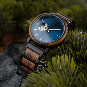 A wristwatch with a blue dial, skeleton movement window, and a band combining wood and black metal, placed on green pine foliage with dark rocky surface in the background.