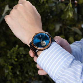 A person wearing a blue dial skeleton wristwatch with wood and black metal band, paired with a light blue striped shirt, against a green foliage background.