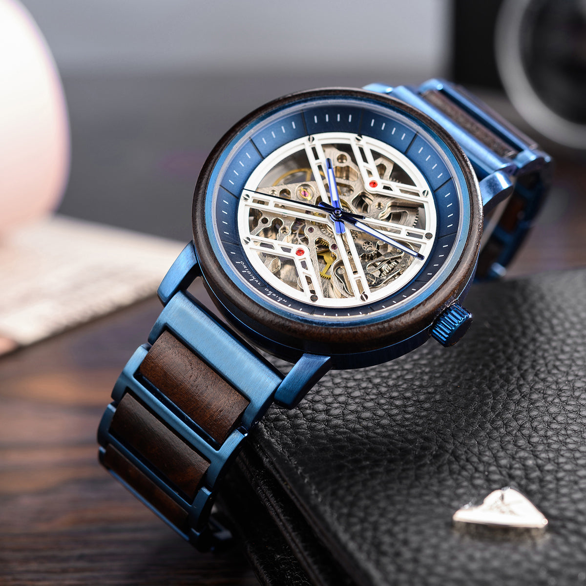 A men's skeleton mechanical watch featuring a blue metal and wooden case and band, showcasing intricate internal mechanisms through a skeleton dial with blue accents, positioned on a black leather surface.
