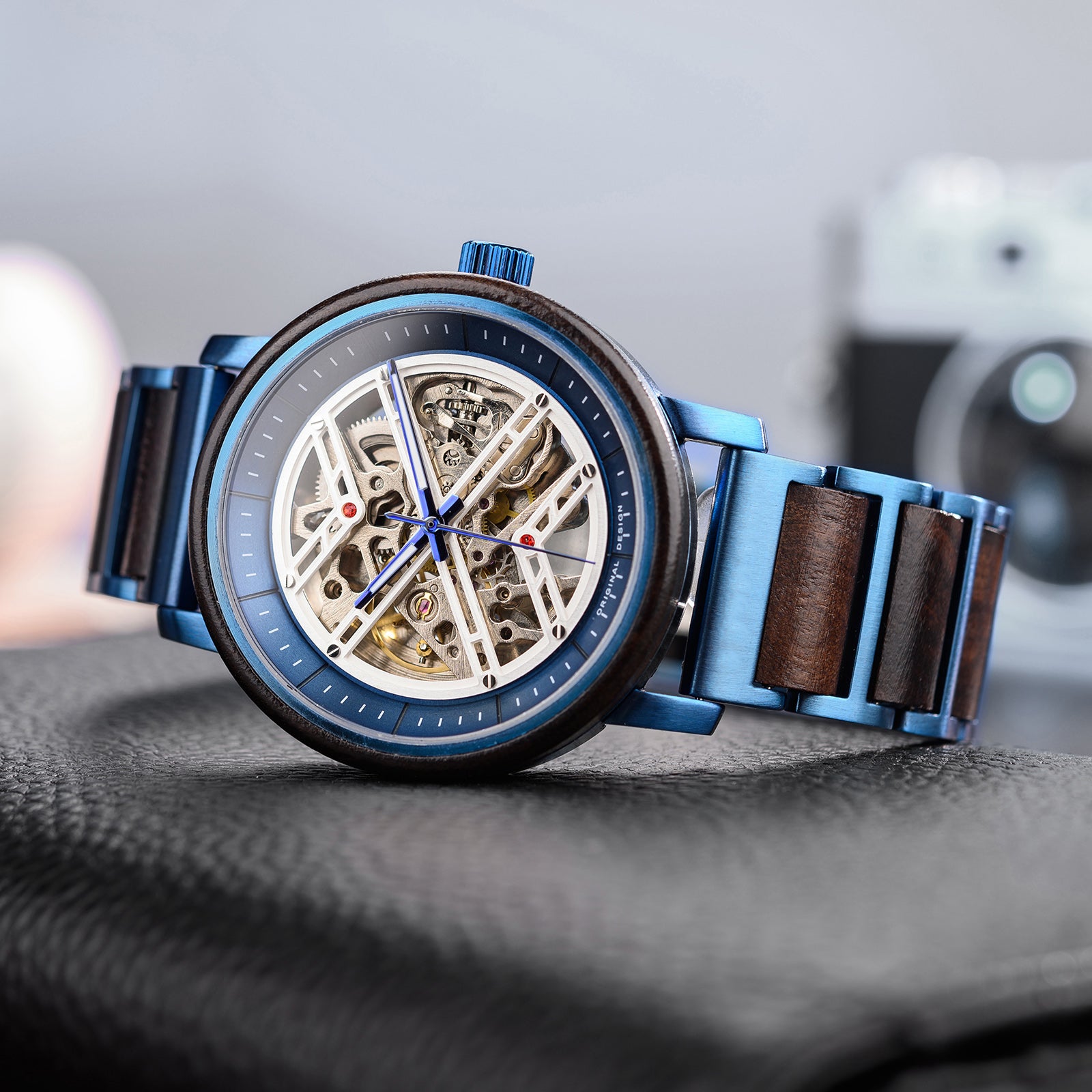 A men's skeleton mechanical watch featuring a blue metal and wooden case and band, showcasing intricate internal mechanisms with red accents through a skeleton dial, positioned on a black leather surface with a blurred background.