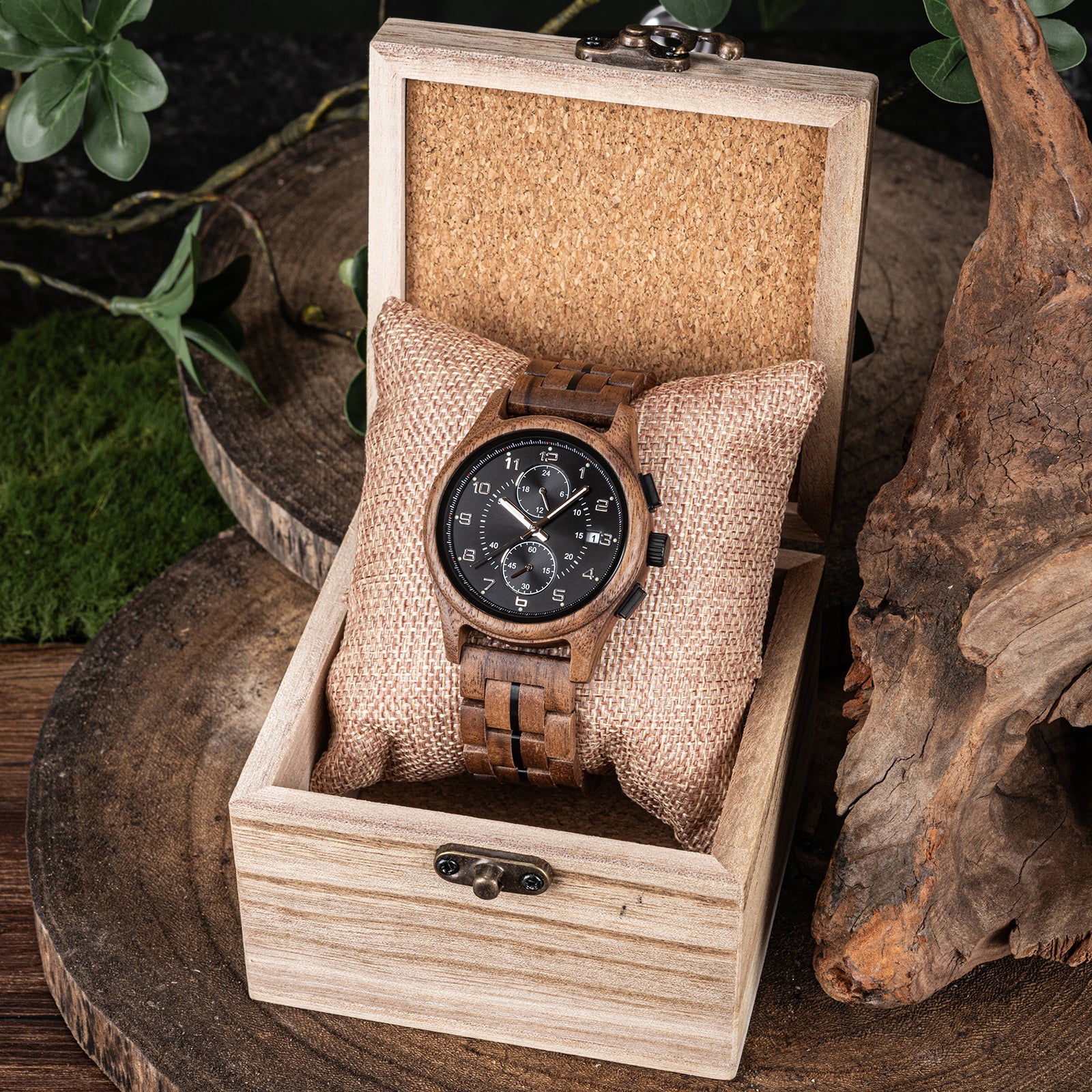 A men's chronograph watch with a brown wooden case and band, placed on a fabric pillow inside a light wooden box, surrounded by natural elements like green foliage, moss, and textured wood.