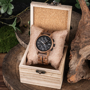 A men's chronograph watch with a brown wooden case and band, placed on a fabric pillow inside a light wooden box, surrounded by natural elements like green foliage, moss, and textured wood.