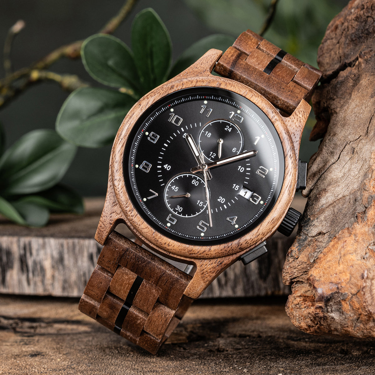 A men's chronograph watch with a brown wooden case and band, featuring a black dial with silver-tone numerals, hands, and sub-dials, positioned on a rustic surface with green foliage and textured wood in the background.