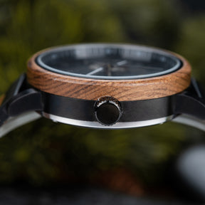 A close-up view of a watch featuring a wooden bezel and a black metal crown, with a blurred green natural background.