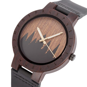 A close-up of a unique dark wooden watch with a runic symbol bezel, forest silhouette dial, black leather band, and gold-tone hands, emphasizing the natural wood texture against a white background.