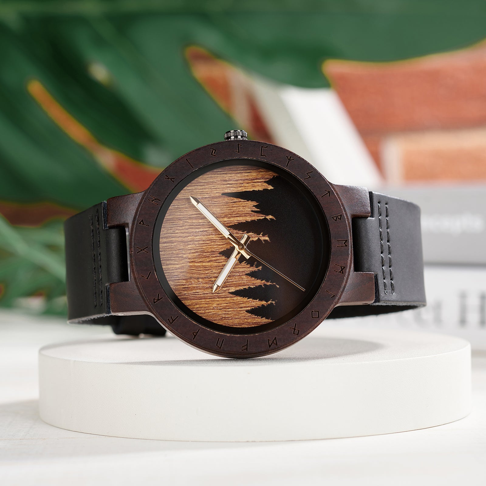 A unique dark wooden watch with a runic symbol bezel, forest silhouette dial, black leather band, and gold-tone hands, displayed on a white platform against a blurred background of green foliage and books.