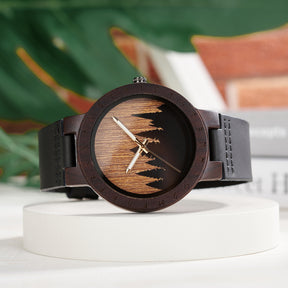 A unique dark wooden watch with a runic symbol bezel, forest silhouette dial, black leather band, and gold-tone hands, displayed on a white platform against a blurred background of green foliage and books.