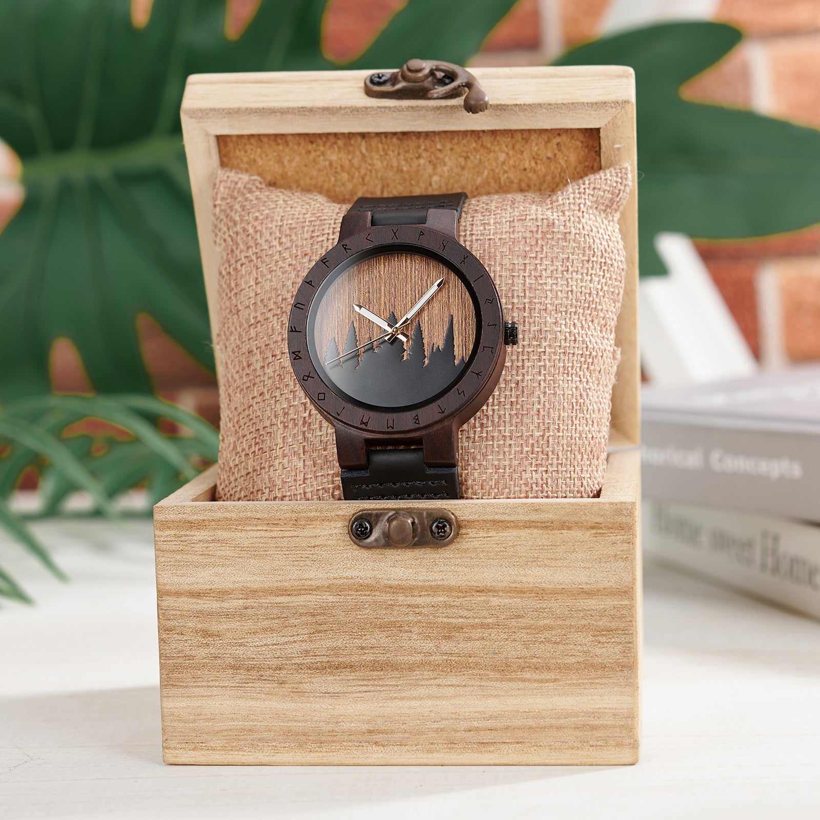 A unique dark wooden watch with a runic symbol bezel, forest silhouette dial, and black leather band, nestled on a burlap pillow inside a wooden box, with a blurred background of green foliage and books.
