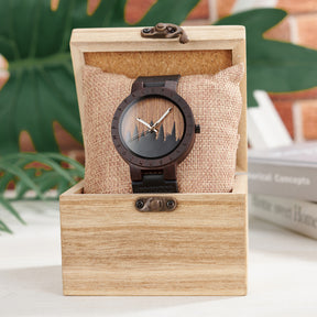 A unique dark wooden watch with a runic symbol bezel, forest silhouette dial, and black leather band, nestled on a burlap pillow inside a wooden box, with a blurred background of green foliage and books.