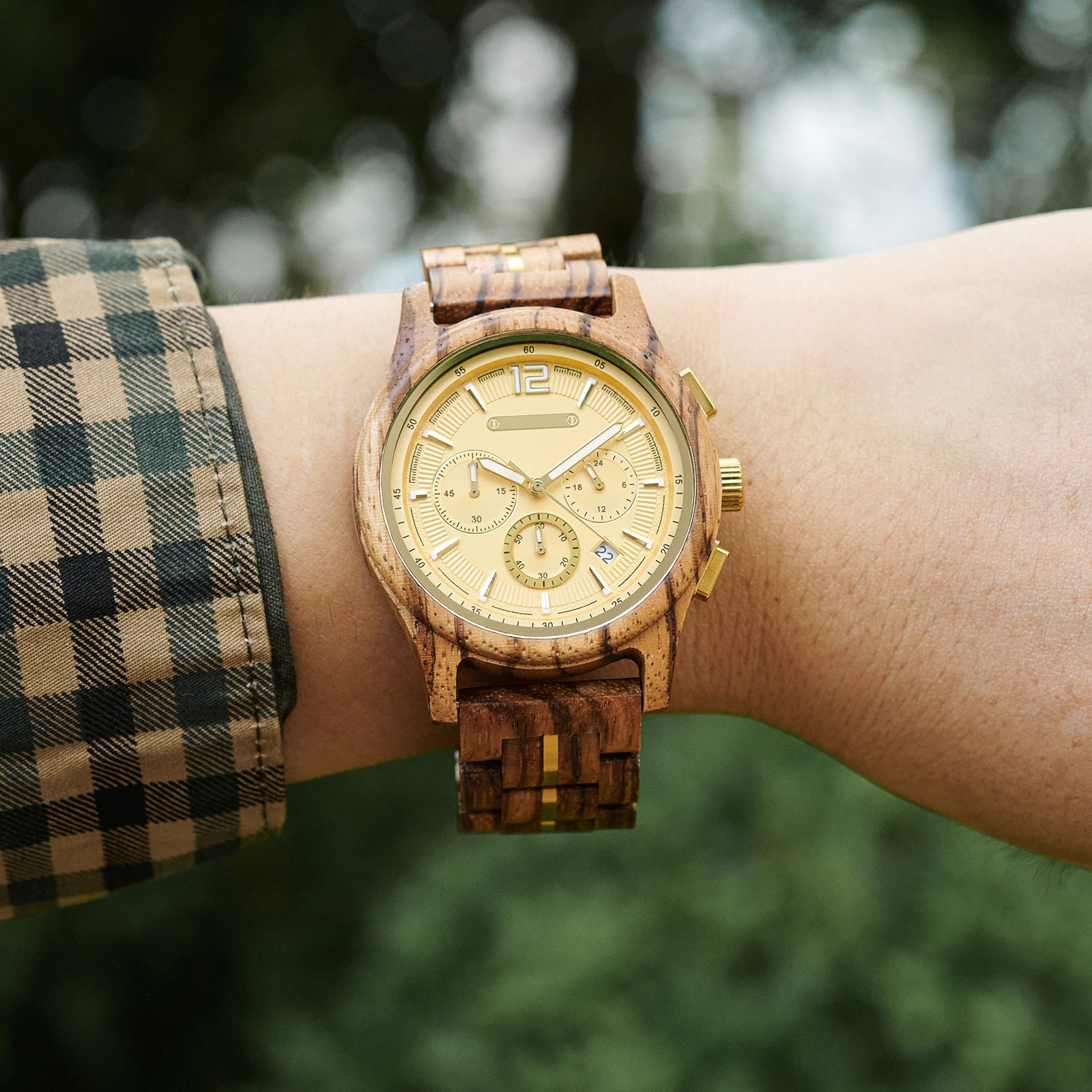 A wooden chronograph watch with a gold dial and gold accents, worn on a wrist 搭配 a plaid shirt, against a blurred green outdoor background.