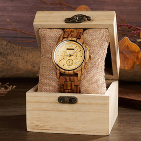 A chronograph watch with a gold dial, wooden case and band (with gold accents), nestled on a burlap pillow inside a wooden box, surrounded by autumnal leaves and a warm wooden background.