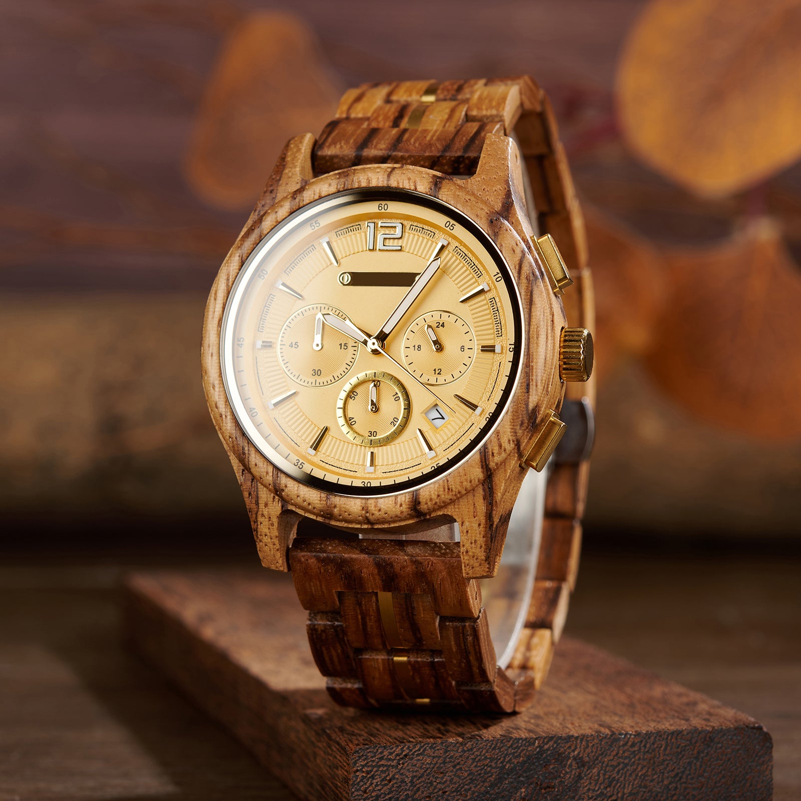 A chronograph watch with a gold dial, wooden case and band (with gold accents), displayed on a wooden block against a warm, blurred background of autumnal leaves.