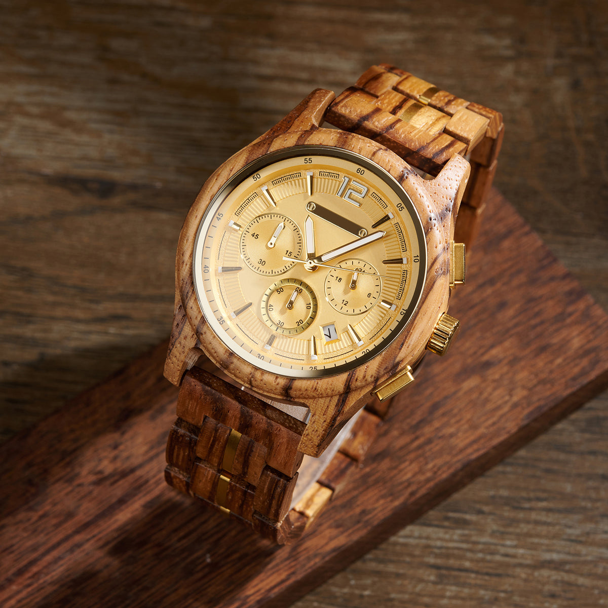 A chronograph watch featuring a gold dial, wooden case and band with gold accents, placed on a wooden block against a textured wood background.