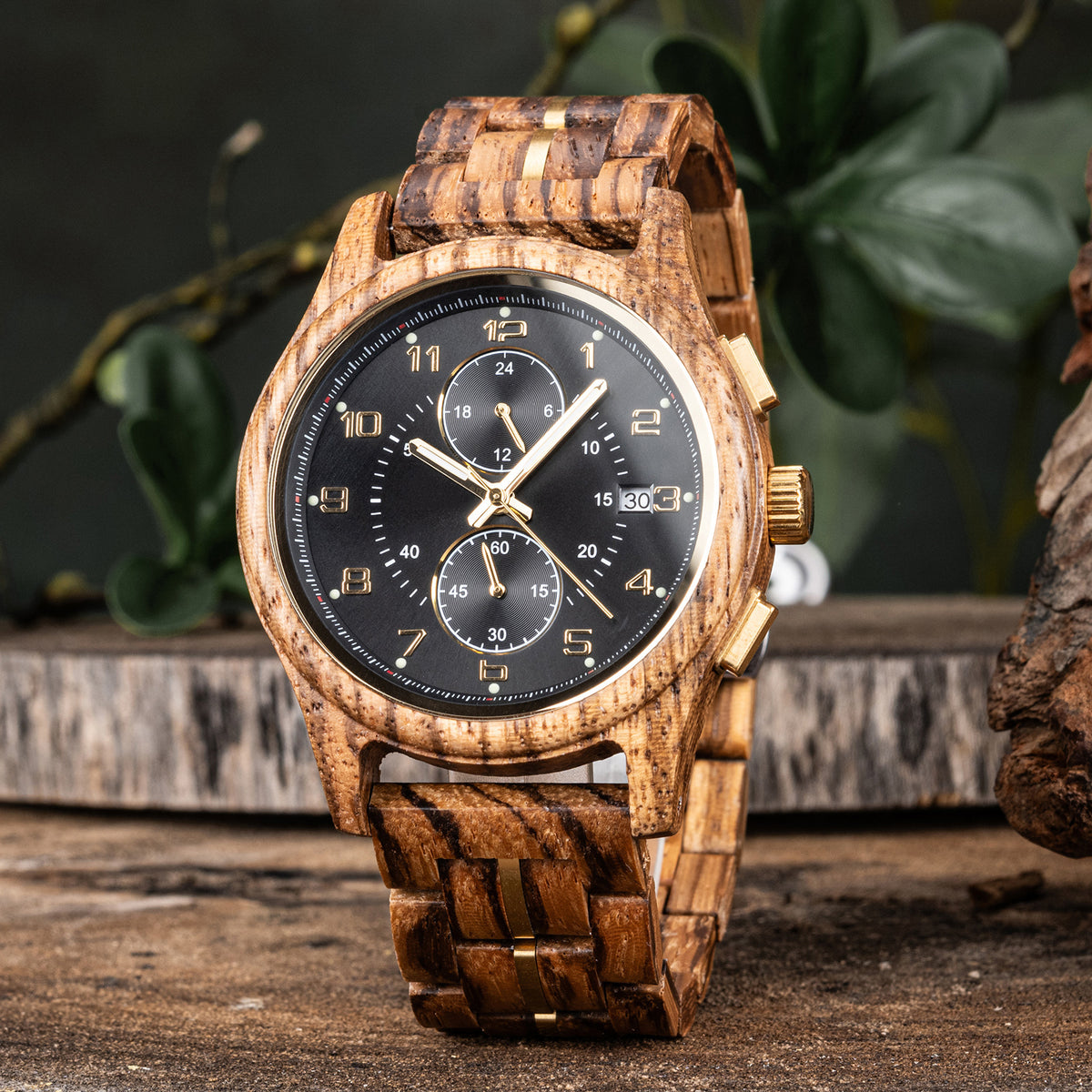 A men's chronograph watch featuring a wooden case and band with gold-tone accents, a black dial with gold numerals, hands, sub-dials, and a date display, set against a rustic background with green foliage.