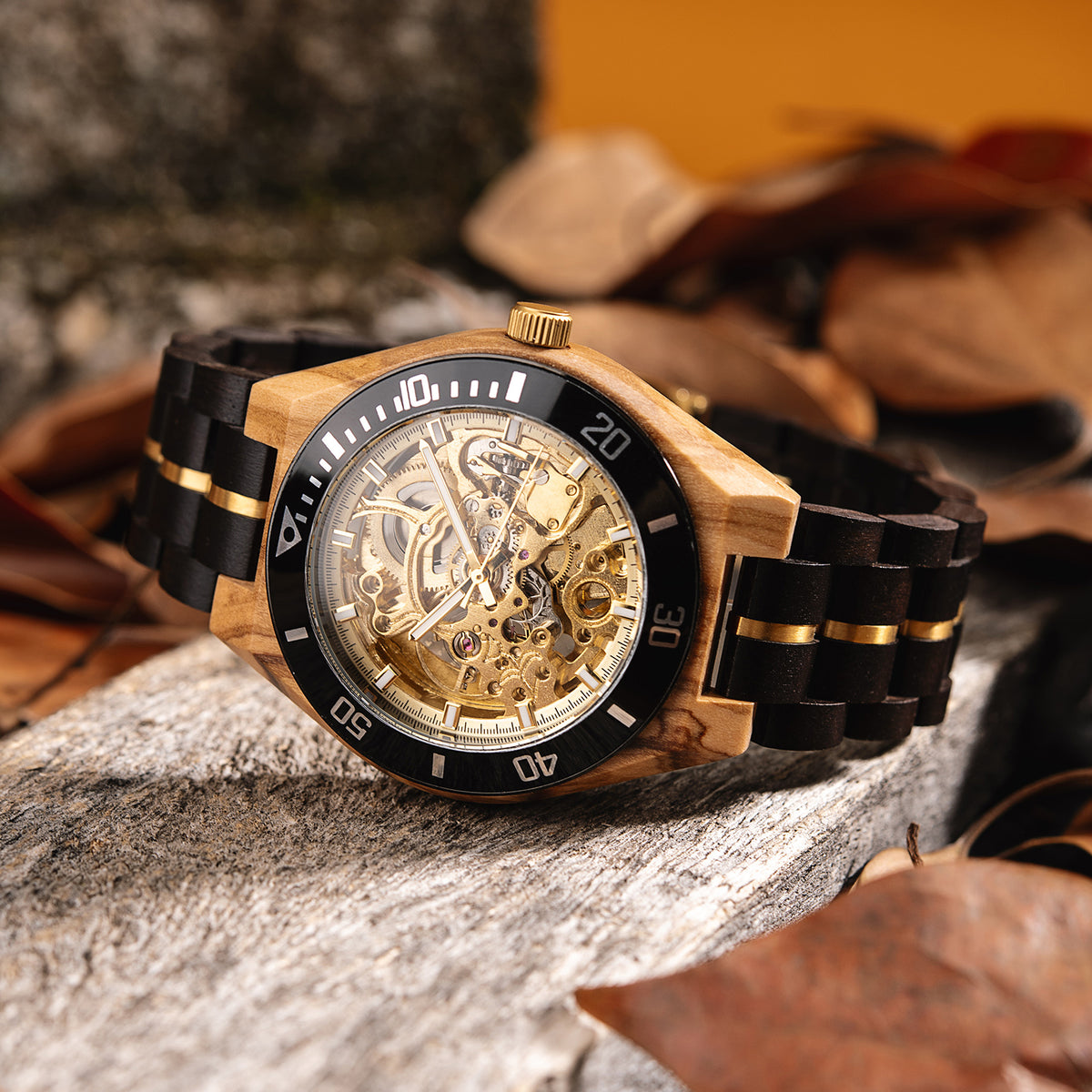 A wooden wristwatch with a gold full skeleton dial, black and gold accented band, placed on a weathered stone surface with autumn leaves in the background.