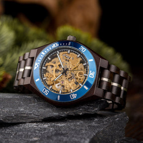 A wooden wristwatch with a gold full skeleton dial, blue bezel, and dark wooden band with silver accents, placed on dark rocks with green pine foliage in the background.