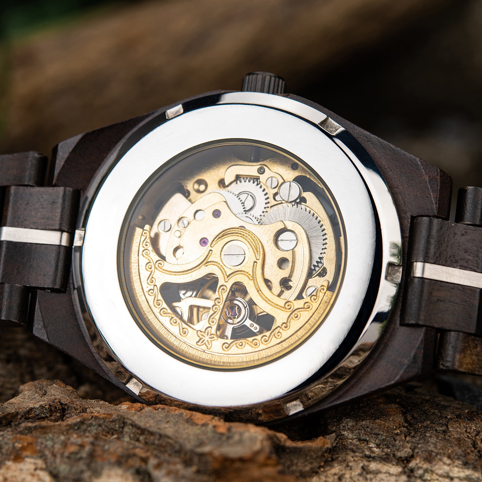 Back view of a wooden wristwatch with an ornate gold skeleton movement, dark wooden band with silver accents, placed on tree bark.