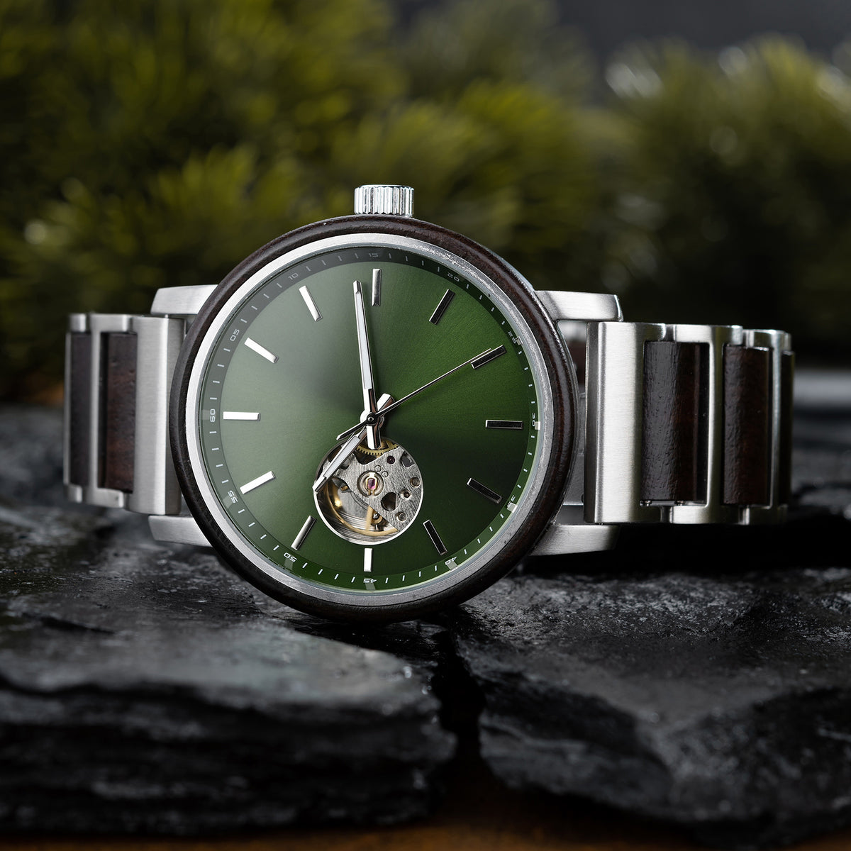 A sophisticated skeleton automatic watch featuring a green dial, wooden bezel, and silver metal with wood combined band, placed on dark rocks with a blurred green foliage background.