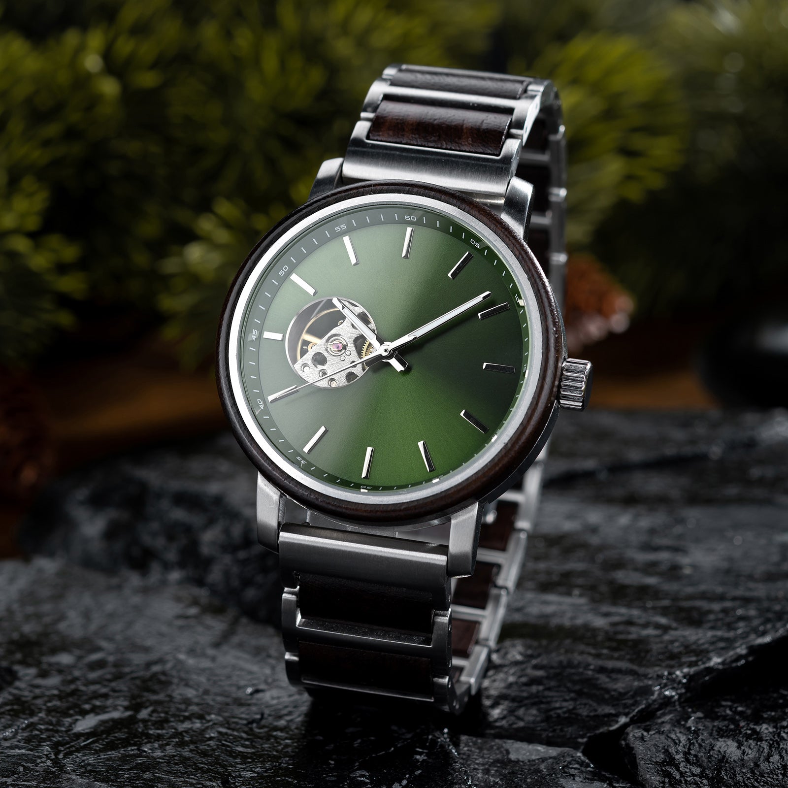 A men's wristwatch with a green dial, skeleton movement window, stainless steel and wood combined band, placed on dark rocks with green foliage in the background.