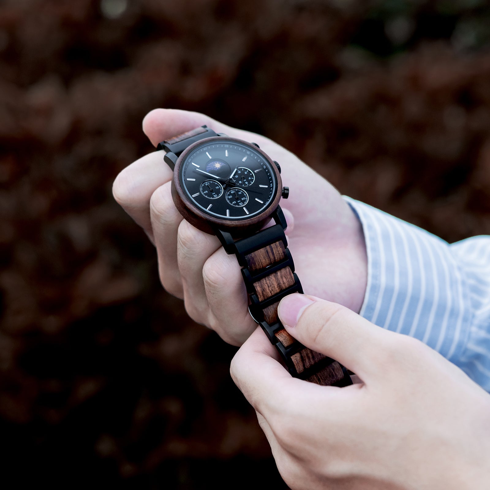 Hands adjusting a stylish black chronograph watch with wood and metal band, featuring a sun-moon phase complication, against a blurred brown natural background.