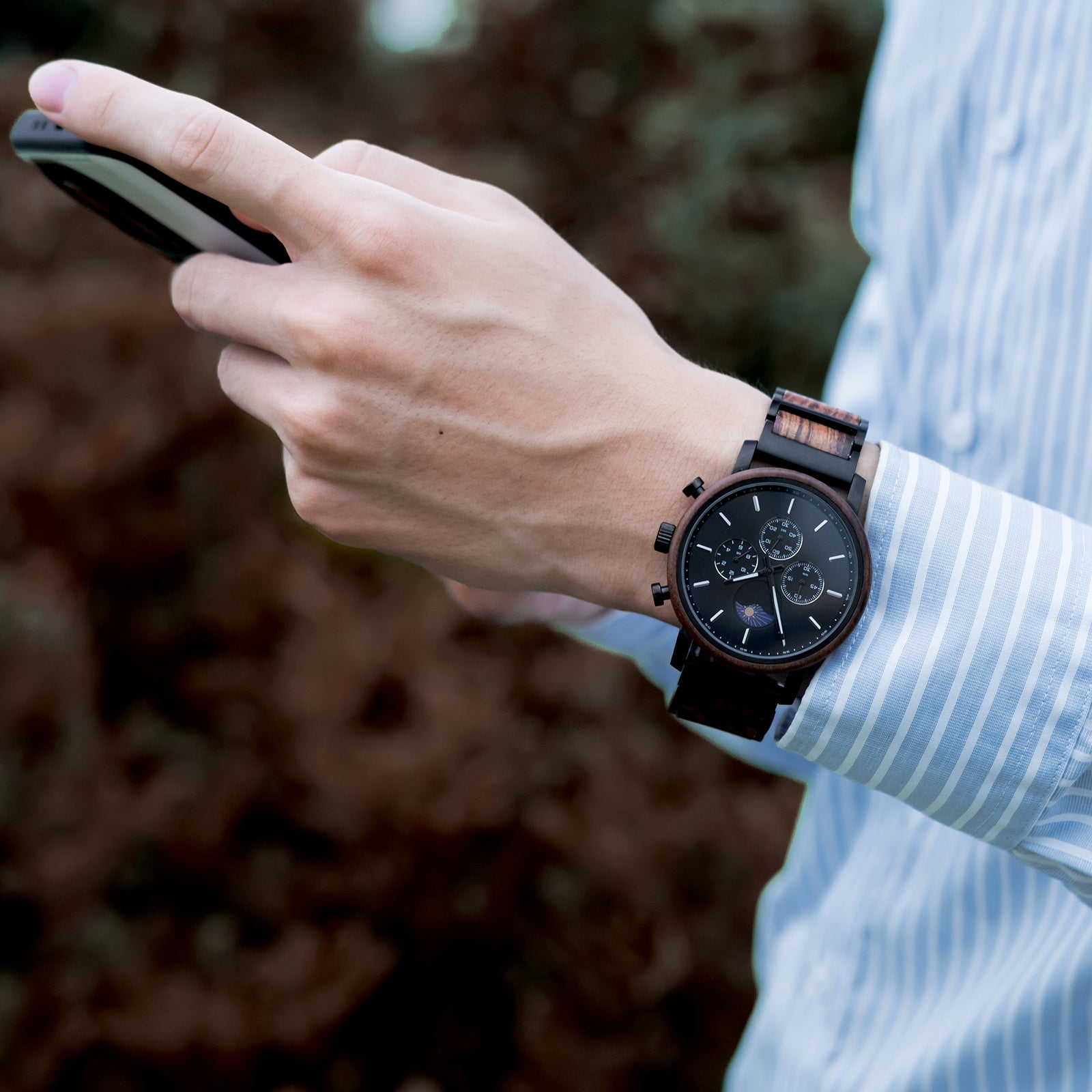 A man in a light blue striped shirt wearing a sophisticated black chronograph watch with wood and metal band (featuring sun-moon phase complication) while using a smartphone, with a blurred natural background.
