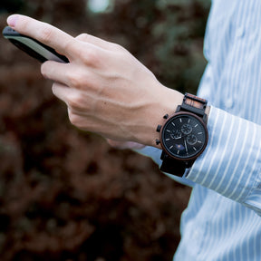 A man in a light blue striped shirt wearing a sophisticated black chronograph watch with wood and metal band (featuring sun-moon phase complication) while using a smartphone, with a blurred natural background.