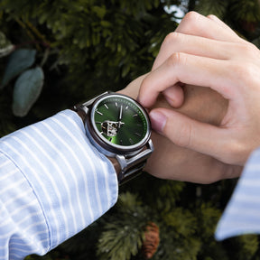 A man in a light blue striped shirt wearing a sophisticated green dial skeleton automatic watch with a wood and metal band, against a backdrop of green foliage.