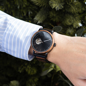A man in a light blue striped shirt wearing a stylish skeleton automatic watch with a wooden bezel and black metal-wood combined band, against a backdrop of green foliage.
