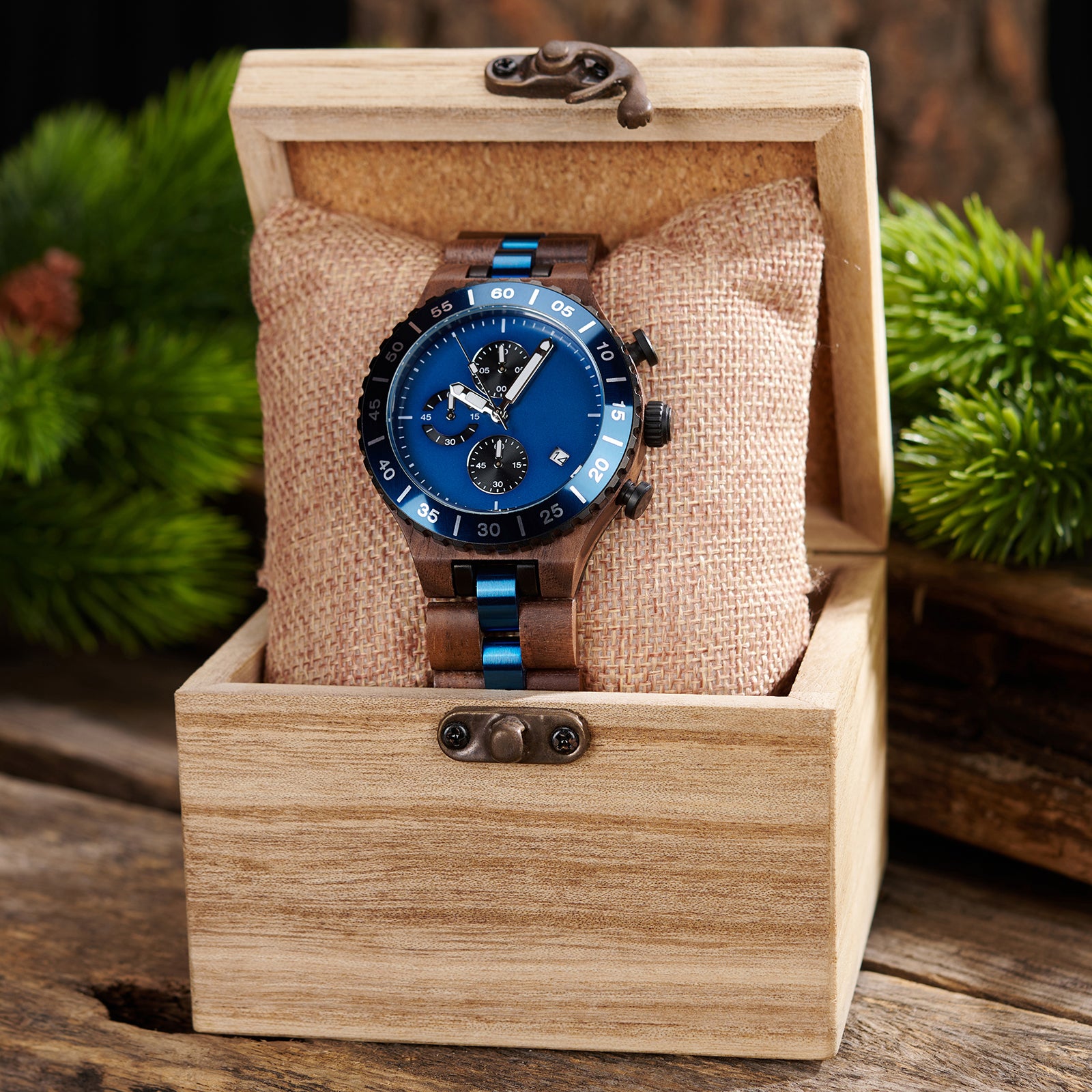 A sophisticated men's walnut wooden chronograph watch with blue accents, presented in a wooden box with a burlap cushion, set against a backdrop of lush greenery to emphasize its natural and refined packaging.