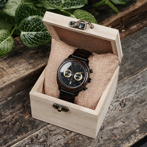 A sophisticated men's wooden chronograph watch with a black dial and gold accents, presented in a wooden box with a burlap cushion, set against a backdrop of lush greenery to emphasize its natural and refined packaging.
