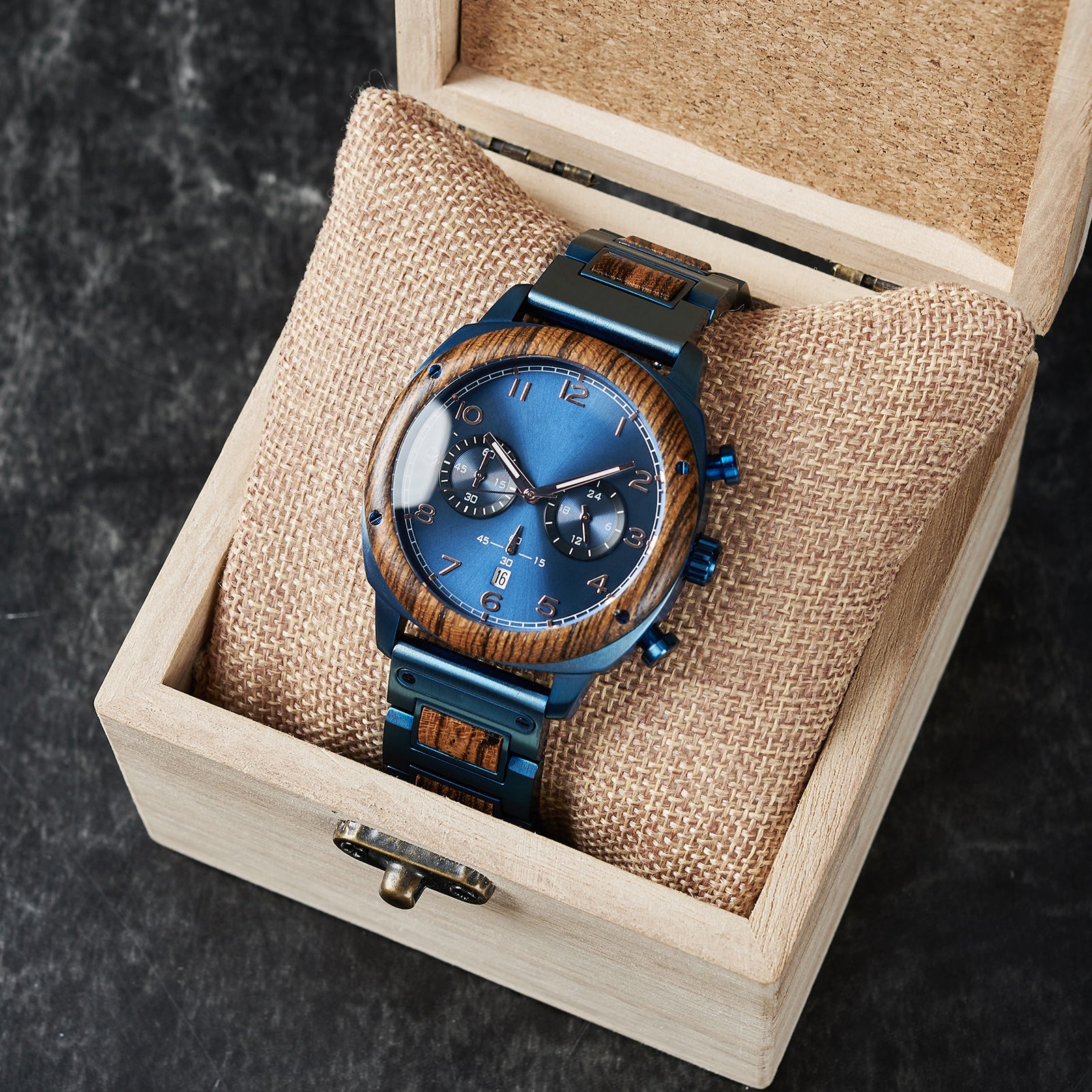 A sophisticated men's wooden chronograph watch with a blue dial, presented in a wooden box with a burlap cushion, set against a dark background to emphasize its refined and elegant packaging.