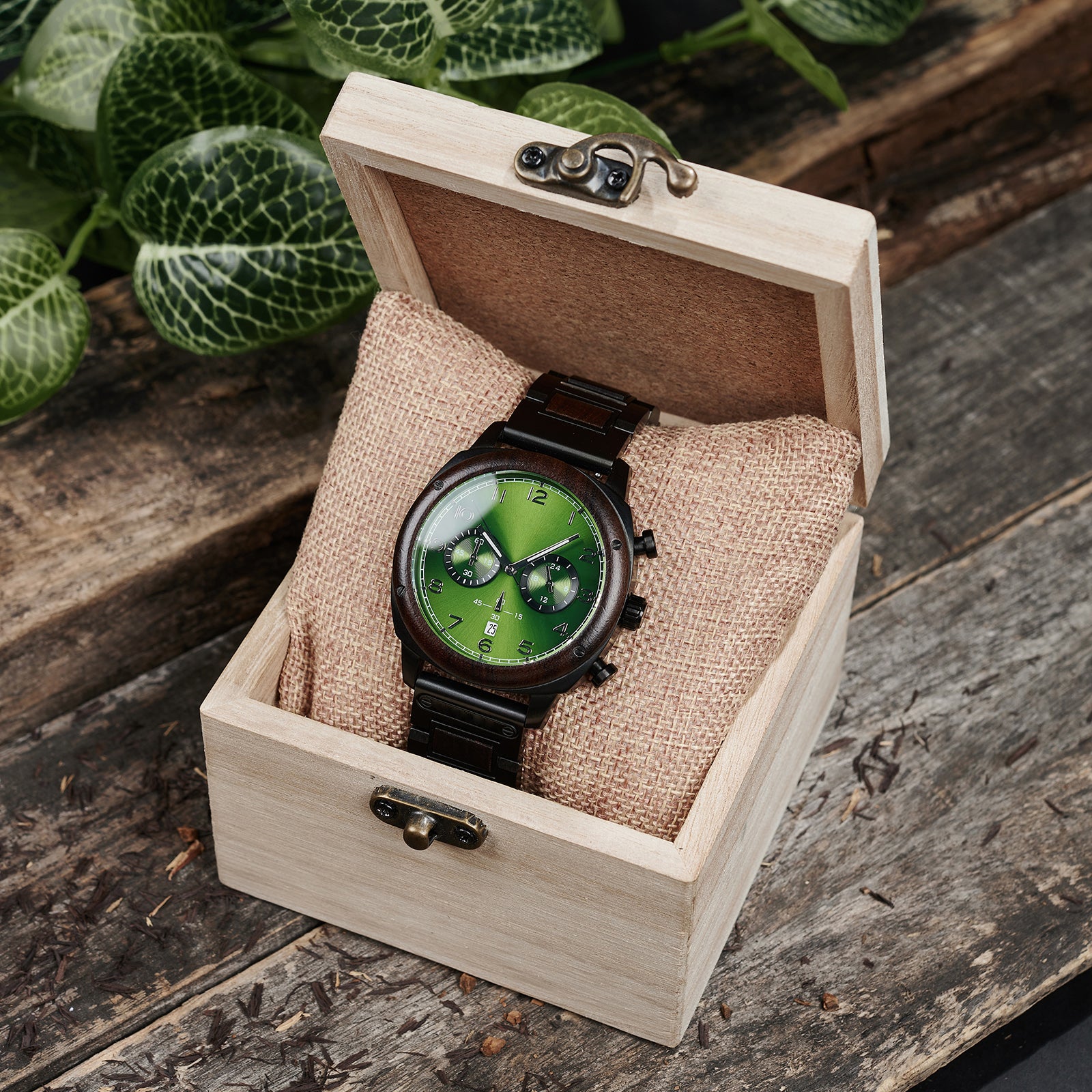 A sophisticated men's wooden chronograph watch with a green dial, presented in a wooden box with a burlap cushion, set against a backdrop of lush greenery to emphasize its natural and refined packaging.