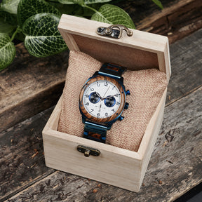 A sophisticated men's wooden chronograph watch with a white dial and blue accents, presented in a wooden box with a burlap cushion, set against a backdrop of lush greenery to emphasize its natural and refined packaging.