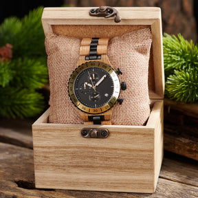 A sophisticated men's wooden chronograph watch with gold and black accents, presented in a wooden box with a burlap cushion, set against a backdrop of lush greenery to emphasize its natural and refined packaging.
