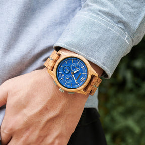 A wooden chronograph watch with a blue dial and olive wood band, worn on a man's wrist 搭配 a light gray shirt, with a blurred green background.
