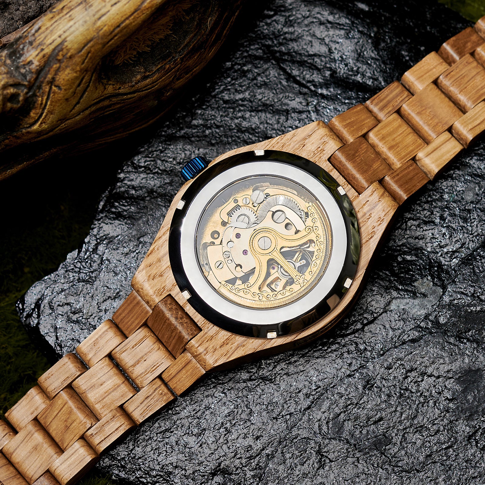 The case back of a men's automatic mechanical watch made of oak wood and black stainless steel, revealing intricate golden mechanical movements with detailed engravings, placed on a textured black rock with a natural wooden element in the background.