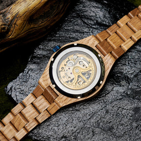 The case back of a men's automatic mechanical watch made of oak wood and black stainless steel, revealing intricate golden mechanical movements with detailed engravings, placed on a textured black rock with a natural wooden element in the background.